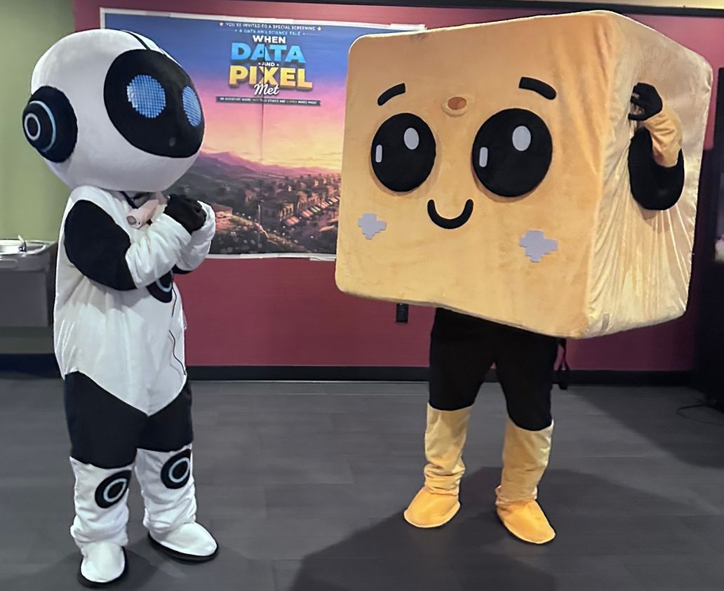 Data and Pixel Costumes Safia Malallah in a plush costume as Data and Lior Shamir in a plush costume of Pixel standing in front of a movie poster for When Data and Pixel Met.